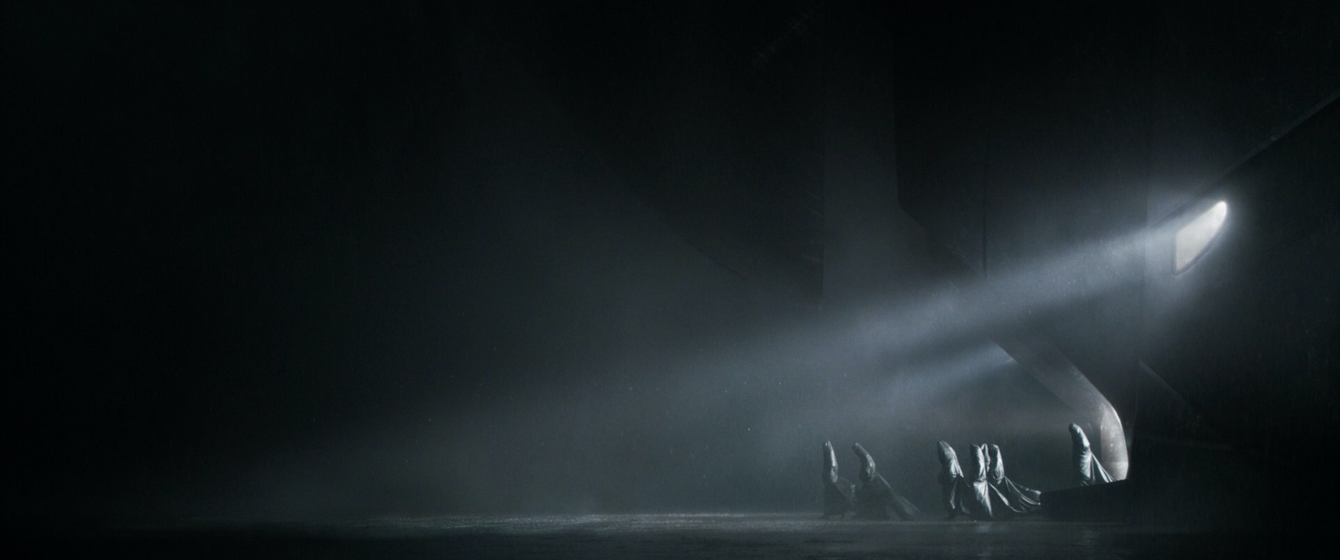 Dune still: a diagonal shaft of light descends from a circular hatch at the upper right; several cloaked figures crouch at its lower-right end; eighty percent of the frame is black.